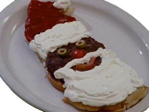 A plated Santa burger with olive eyes, cherry tomato nose, tomato slice mouth, cream cheese beard, and ketchup hat.