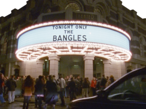 A crowd of people mills around outside a brightly lit theater. The marquee reads "Tonight Only The Bangles."