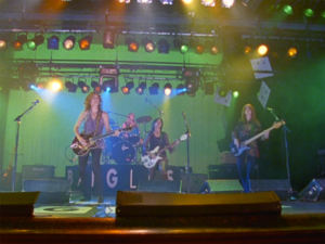 The Bangles, three women with guitars and one on drums, perform on a stage with a green backdrop under rows of lights.